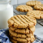 A stack of golden-brown peanut butter cookies with a crisscross pattern on top, sitting on a blue and white checkered cloth. A jar of milk and a cooling rack with more cookies are in the background